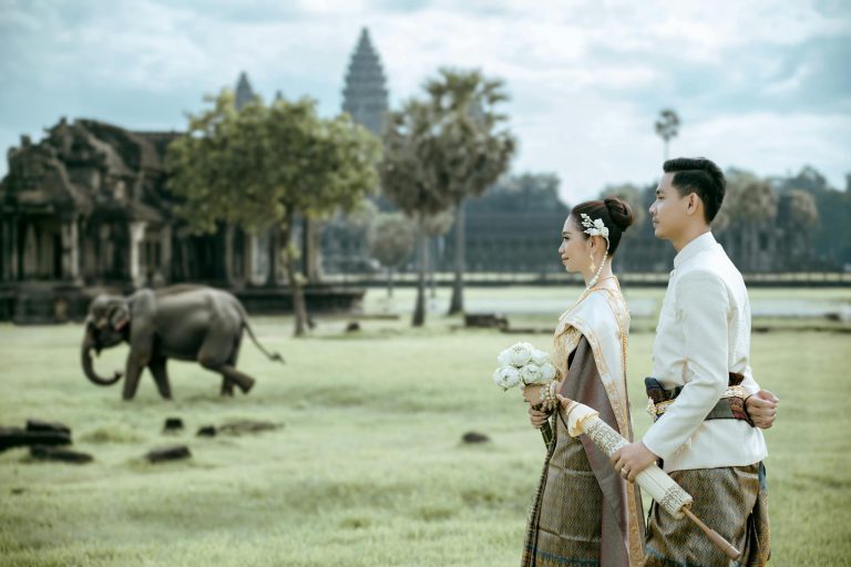 Elegant Cambodian couple in traditional attire at Angkor Wat with an elephant in the background.